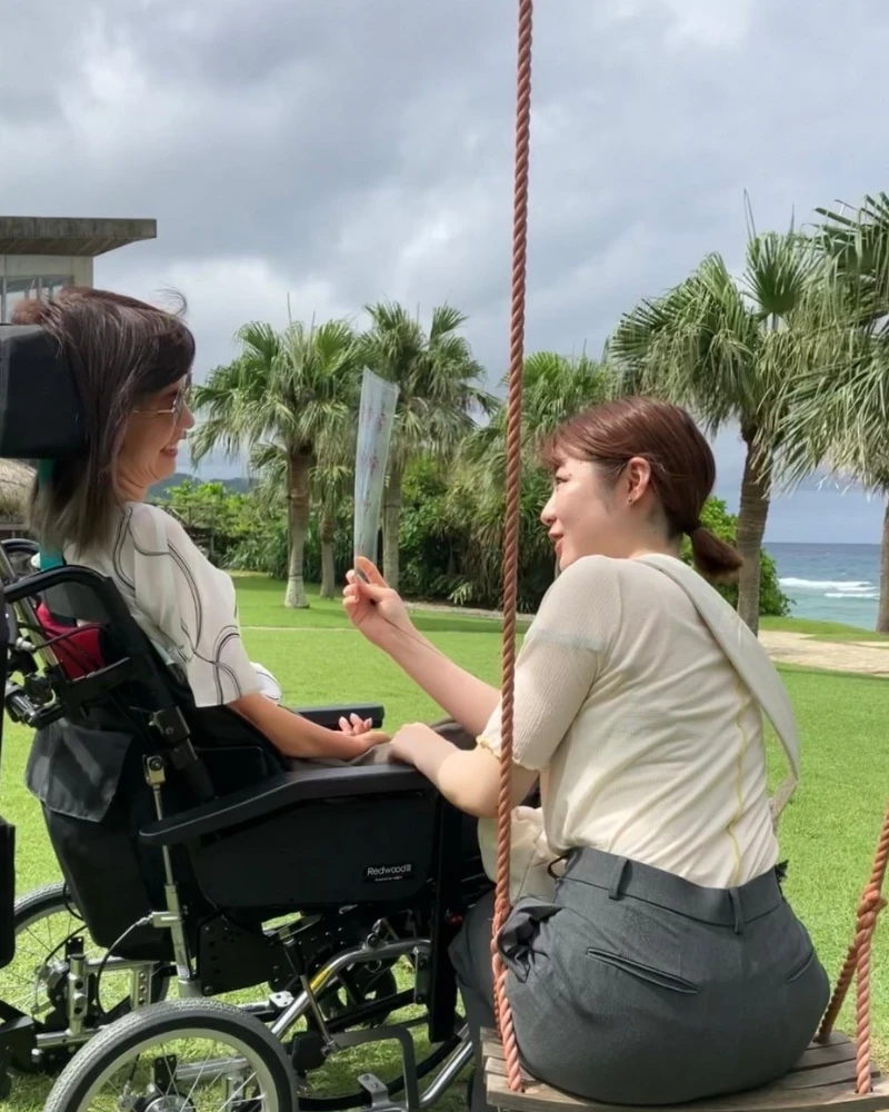 南国の海をバックに車椅子の女性利用者とブランコに乗りながら談笑する介護ヘルパー