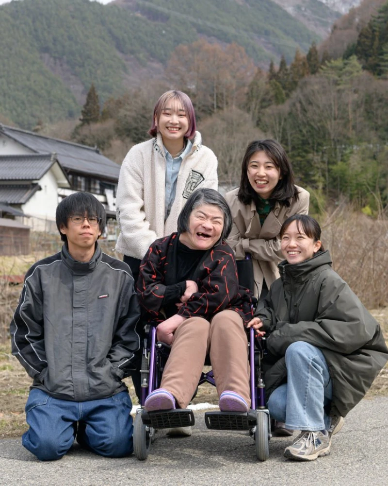 山々を背景に車椅子の利用者を中心に笑顔で記念撮影をする介護スタッフ一同