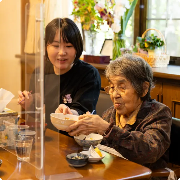 アクリル板越しに高齢女性の食事を介助するスタッフ